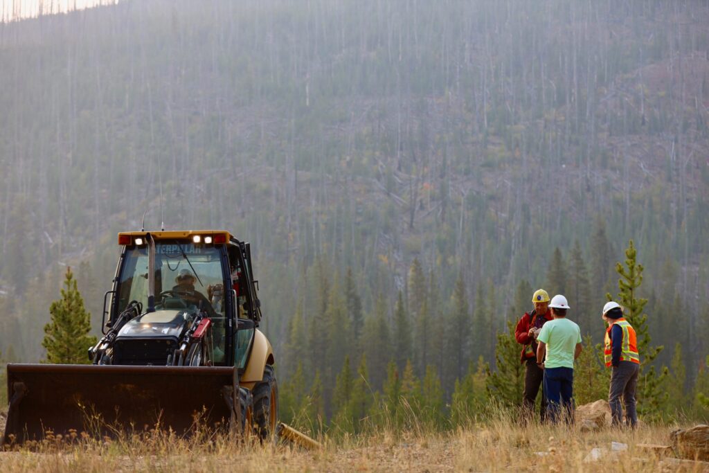 Workers in hard hats and safety vests talking beside a backhoe at the Stibnite Gold Project site